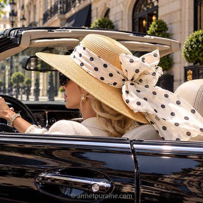 A woman in a black convertible car wearing a large straw sun hat with a wide beige polka dot scarf in silk chiffon, by Anne Touraine Paris. The photo is taken from behind, showing her profile and the car door.