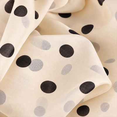 A close-up of a lightweight cream-colored fabric with a polka dot pattern in black and soft beige, shown in soft folds. This is from the beige polka dot scarves by Anne Touraine Paris