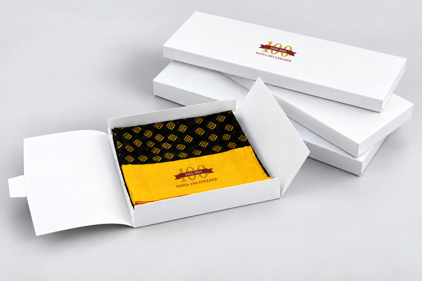 A white gift box set with an open front box revealing a folded black and gold patterned fabric item with a bright yellow band labeled '100 Years Celebrating' and 'Safely Handled.'.
