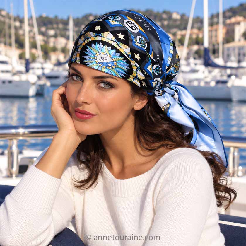 A beautiful brunette is sitting with a marina behind her. She wears on her head, knotted like a pirat, an Anne Touraine Paris French silk scarf "astrology" blue.