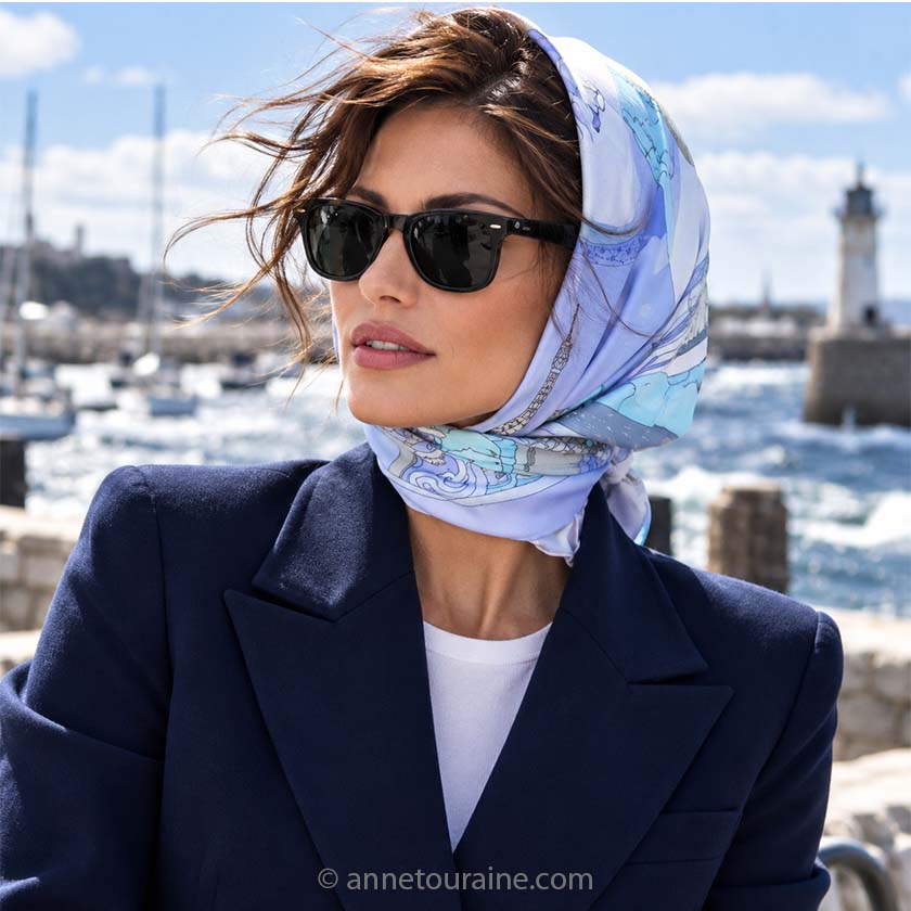 Beautiful fark hair woman with sunglasses, standing nearby a typical French Britany harbor, is wearing on her head an Anne Touraine Paris French silk scarf "Paris", blue.