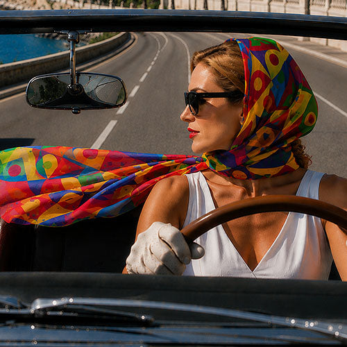 A woman driving a luxury convertible car on a coastal riviera road, wearing a huge vibrant multicolor geometric patterned headscarf in silk chiffon, rayban sunglasses, and a white sleeveless top and gloves.