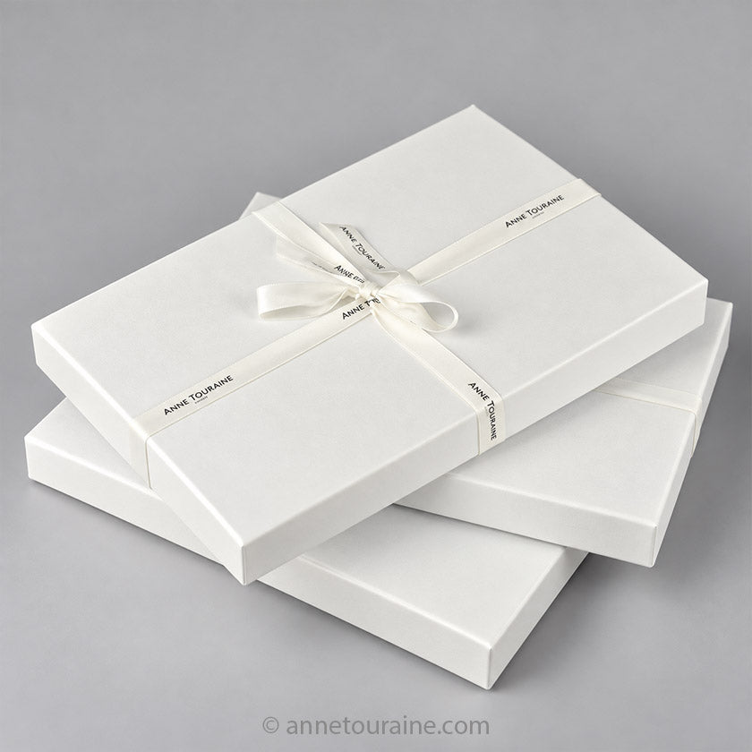 Three white rectangular Anne Touraine Paris gift boxes stacked on a light gray background, with the top box tied in a cross pattern with a white satin ribbon printed with the brand name 'Anne Touraine'.