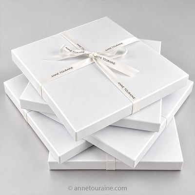 A stack of white rectangular Anne Touraine Paris signature gift boxes tied with white signature cotton ribbons printed with the brand name 'Anne Touraine' on a light gray background.